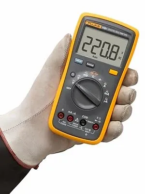 Gloved hand holding a Fluke digital multimeter displaying 220.8 volts on a white background